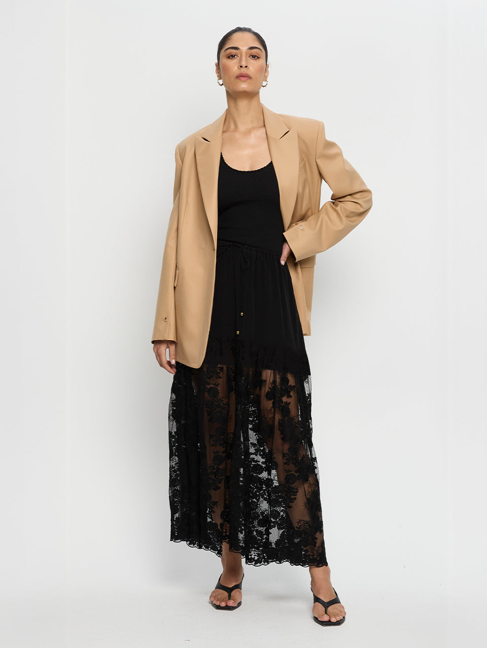 Woman wearing a beige blazer over a black dress with a lace overlay on a white background