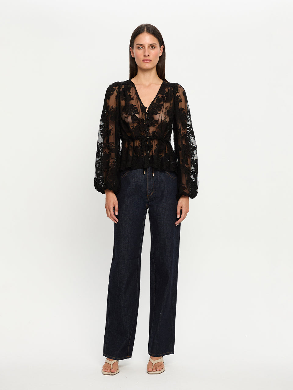 Woman wearing a sheer black blouse with a V-neckline and dark pants on a white background