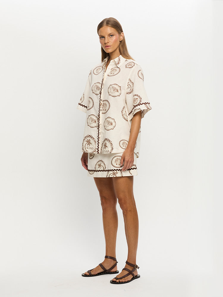 A model wearing a short-sleeved button-down shirt with a pattern of shells and coastal-inspired stamps on an ivory base, paired with a short skirt and sandals.