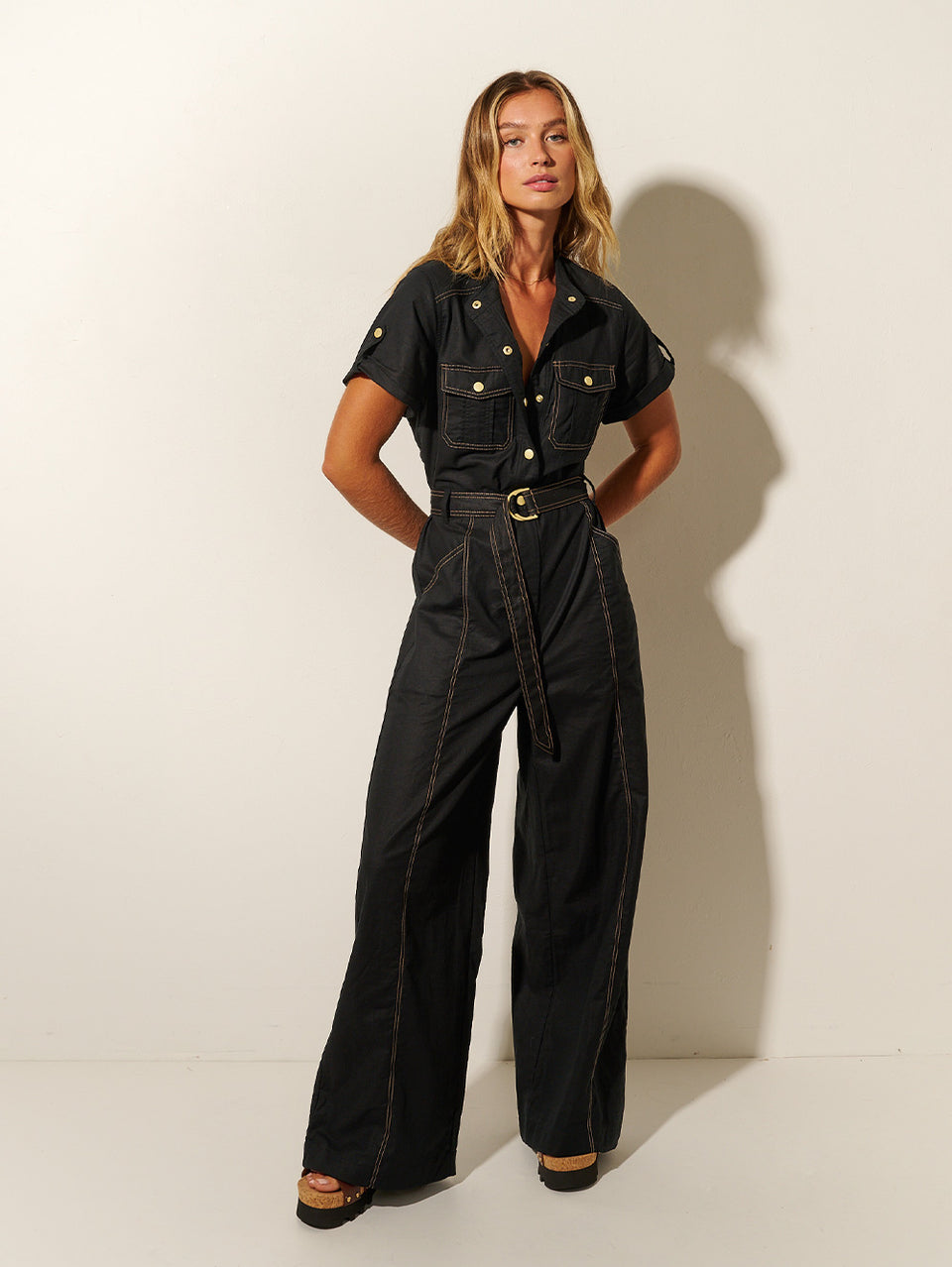 Studio model wears the KIVARI Ebony Jumpsuit: a black linen jumpsuit with gold buttons, topstitch details, a button-front with signature hardware and belt.