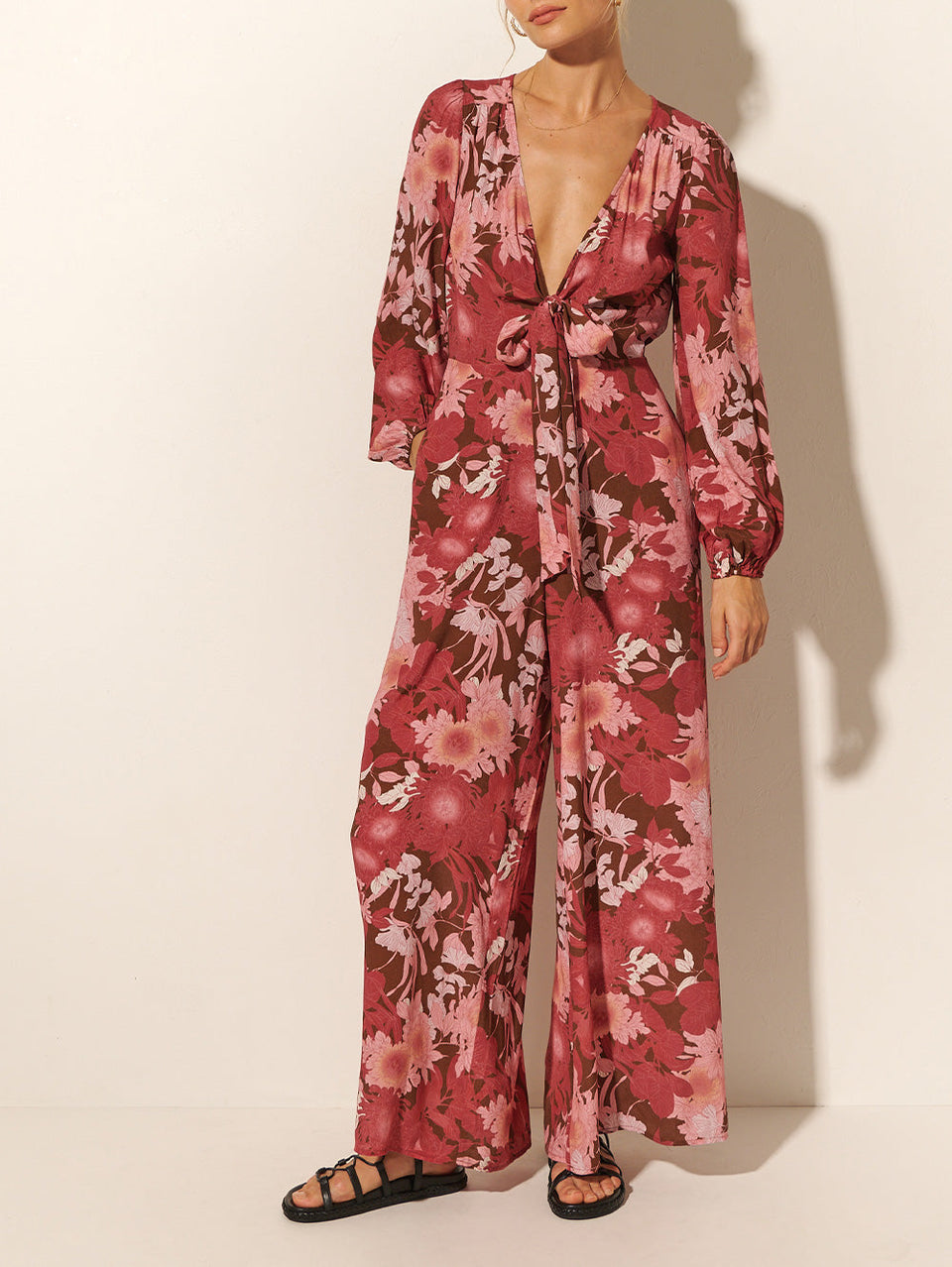 Close-up: Studio model wears KIVARI Hacienda Jumpsuit: A red, pink and brown floral jumpsuit with ribbon tie front and long sleeves, crafted from sustainable LENZING Viscose Crepe.