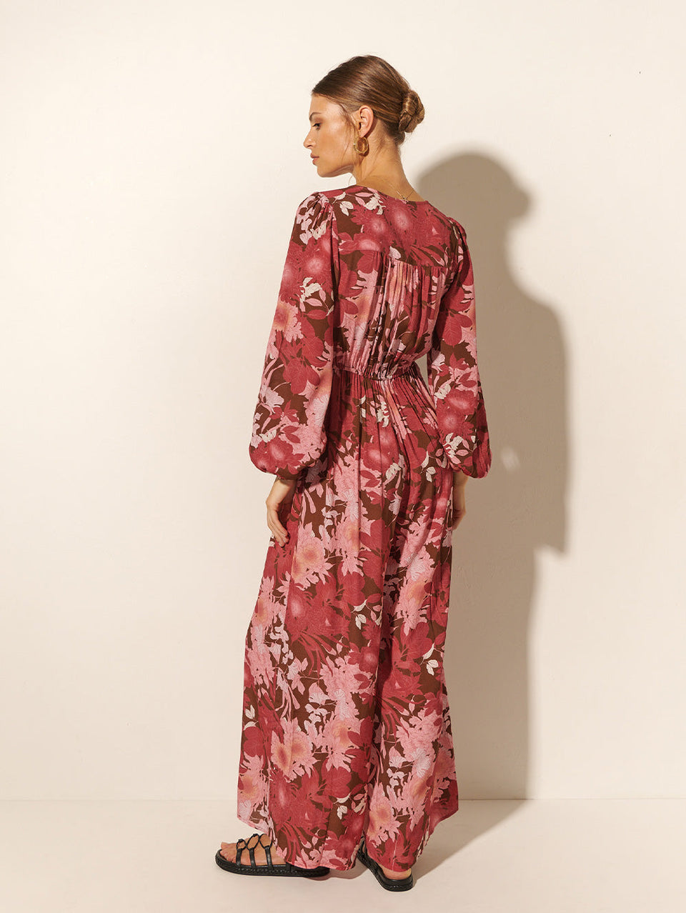 Back shot: Studio model wears KIVARI Hacienda Jumpsuit: A red, pink and brown floral jumpsuit with ribbon tie front and long sleeves, crafted from sustainable LENZING Viscose Crepe.