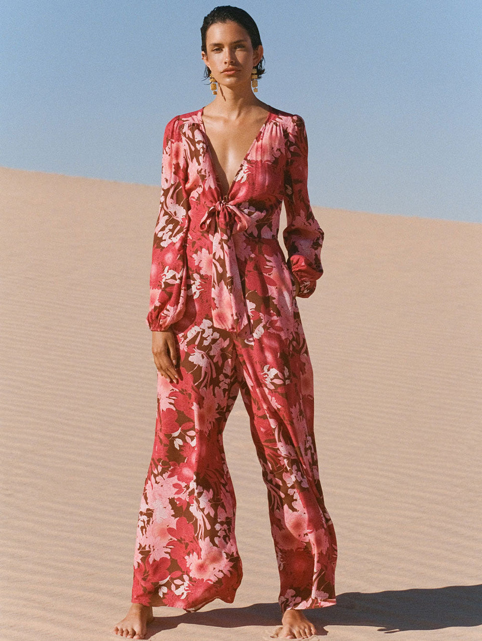 Campaign model in desert wears KIVARI Hacienda Jumpsuit: A red, pink and brown floral jumpsuit with ribbon tie front and long sleeves, crafted from sustainable LENZING Viscose Crepe.