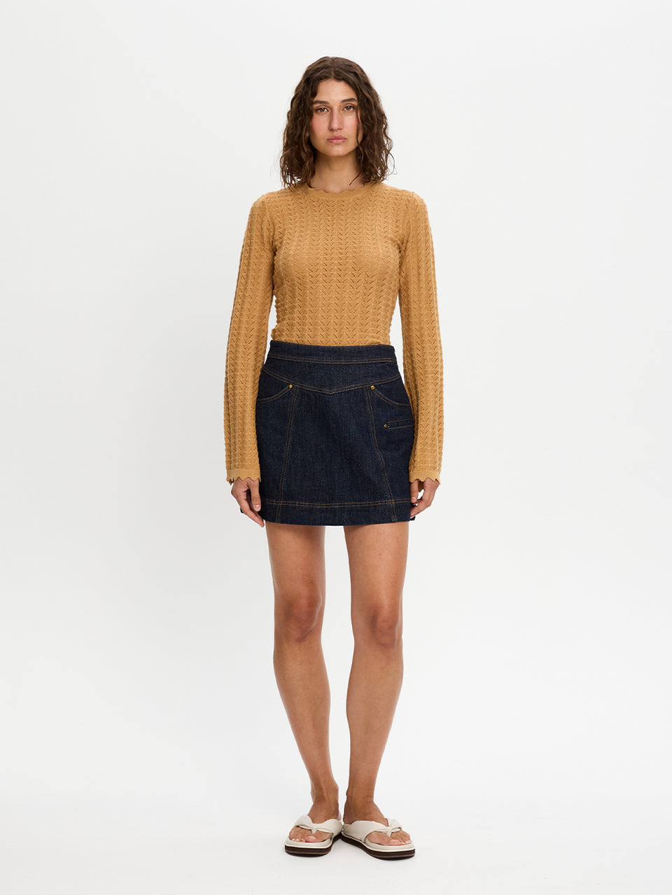 Woman wearing a mustard yellow sweater and dark denim skirt on a white background
