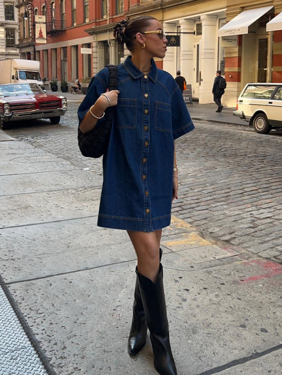 Woman in a denim dress and black boots standing on a city street.