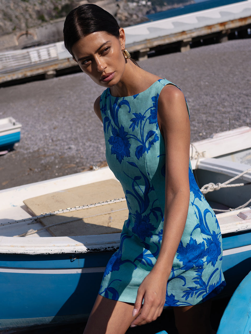 Woman in a blue floral dress standing near boats on a dock.