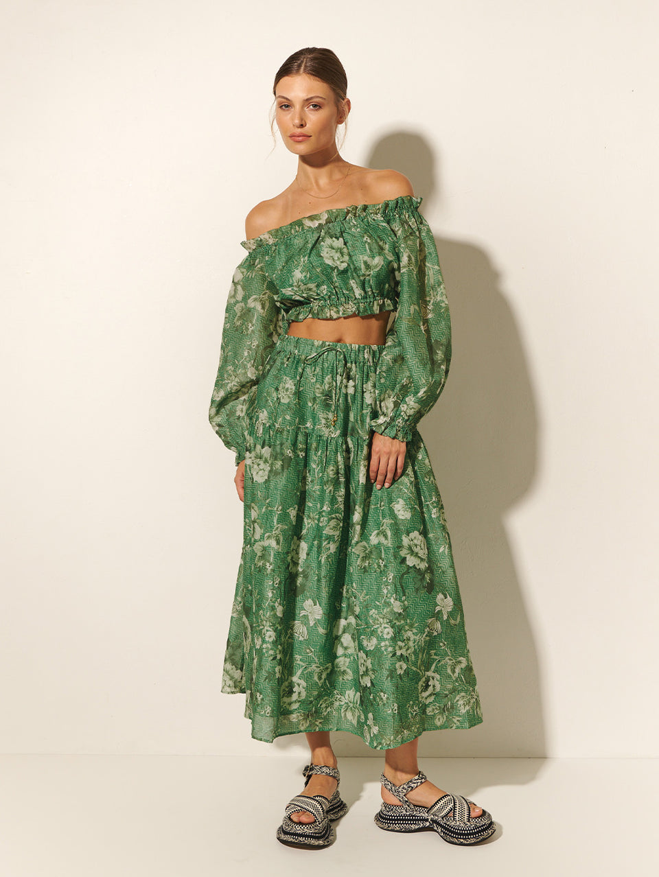 Studio model wears KIVARI Khalo Crop Top: A green floral off-the-shoulder top with full-length blouson sleeves and elasticated bust, waist and sleeve details.