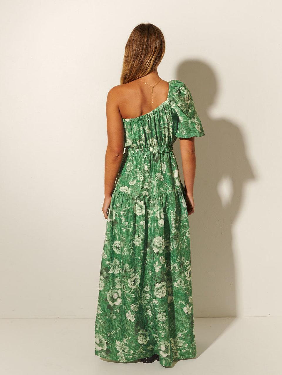 Back shot: Studio model wears KIVARI Khalo Maxi Dress: A green floral one-shoulder dress with a short sleeve, elasticated front and waist, and tiered skirt.