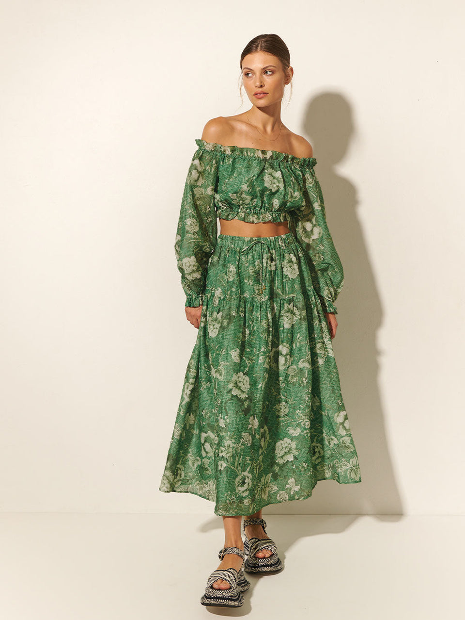 Studio model wears KIVARI Khalo Midi Skirt: A green floral midi skirt featuring an elasticated drawstring waist with gold bead ends and a gathered tiered skirt.