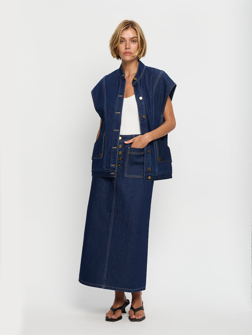 A woman is standing, wearing a long denim midi skirt with contrast topstitch detailing and patch pockets, complete with gold hardware.