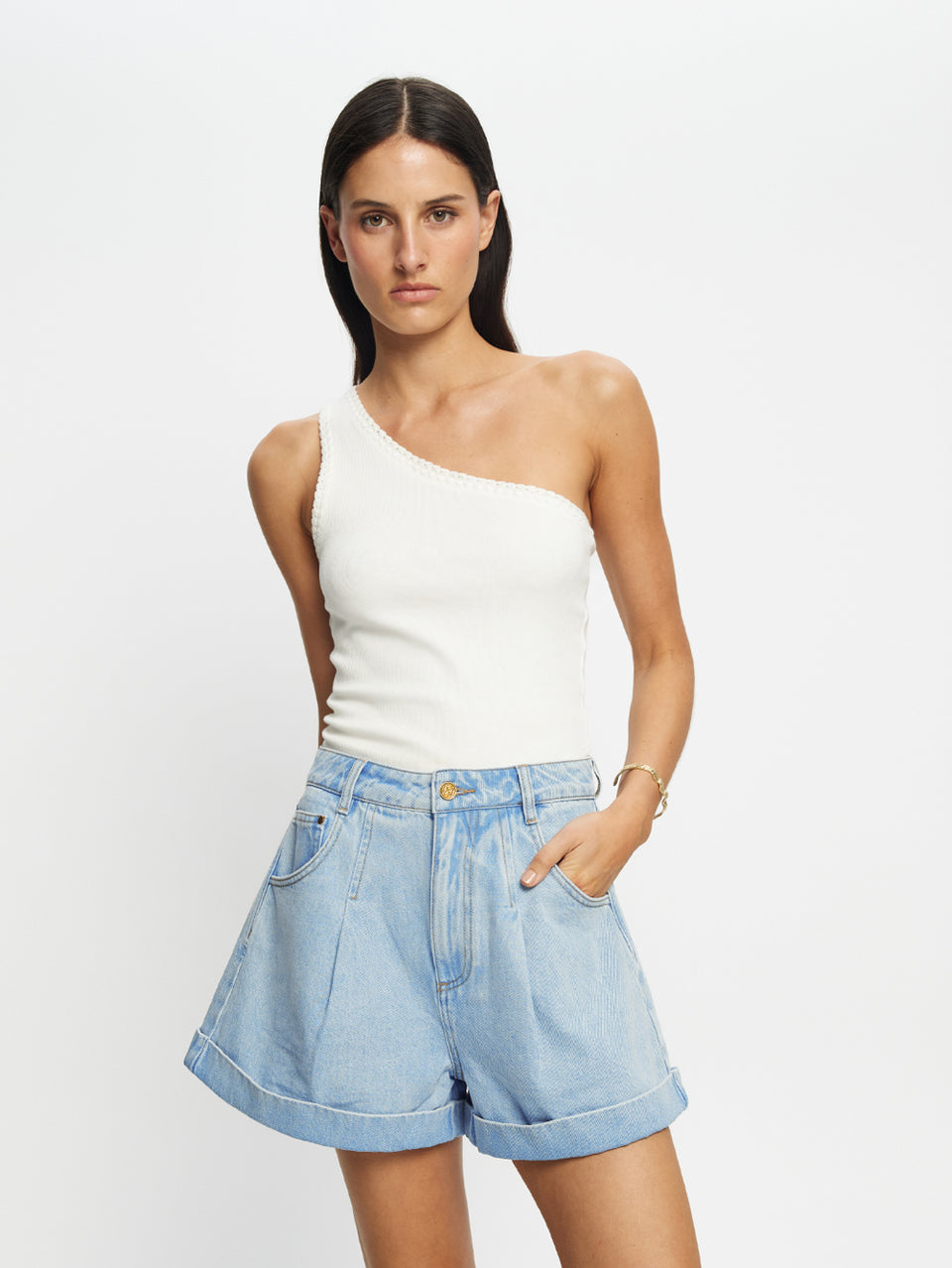 Woman wearing a white one-shoulder top and light blue denim shorts on a plain background