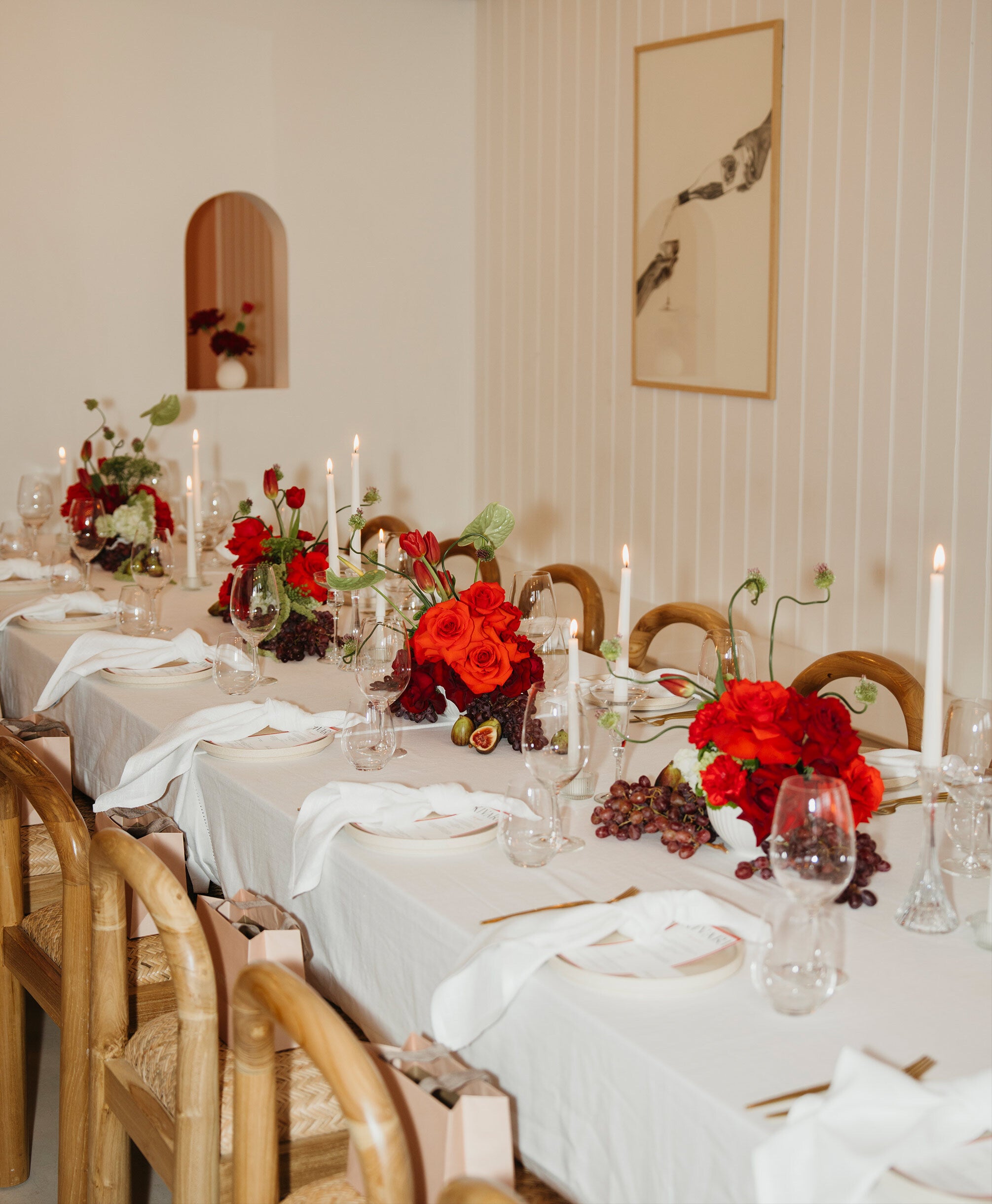 Elegant dining table set with red floral arrangements, candles, and white tablecloth in a room with wooden chairs and wall art.
