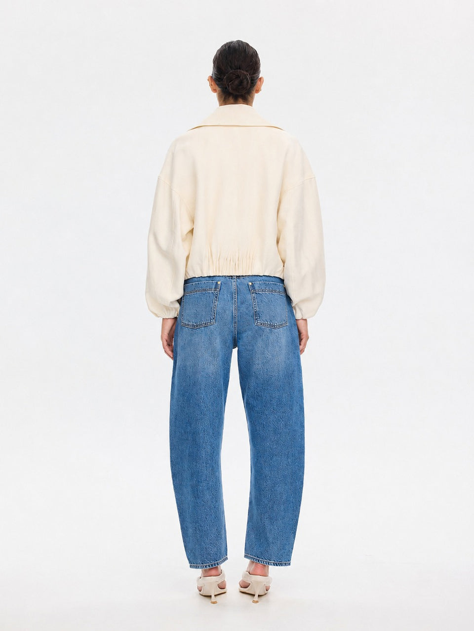 A model viewed from the back wearing a cream-colored cropped jacket and wide-leg blue jeans, standing against a plain white studio background.
