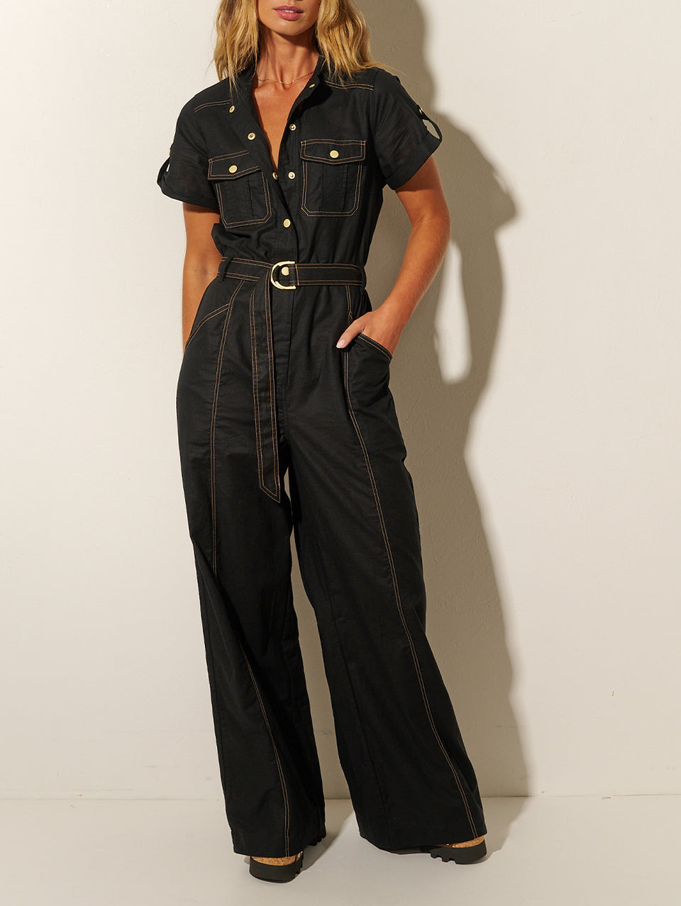 Close up: Studio model wears the KIVARI Ebony Jumpsuit: a black linen jumpsuit with gold buttons, topstitch details, a button-front with signature hardware and belt.