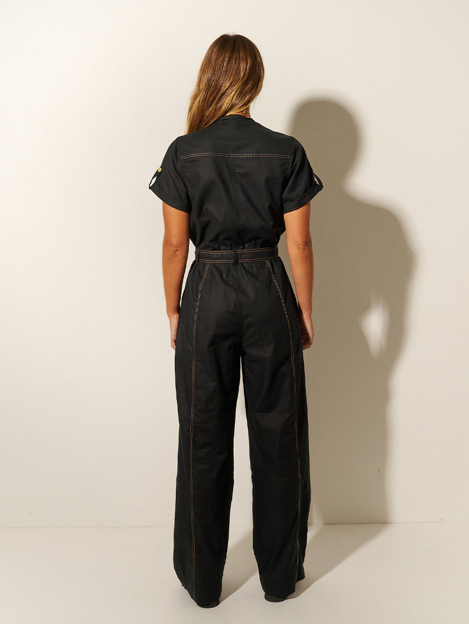 Back shot: Studio model wears the KIVARI Ebony Jumpsuit: a black linen jumpsuit with gold buttons, topstitch details, a button-front with signature hardware and belt.