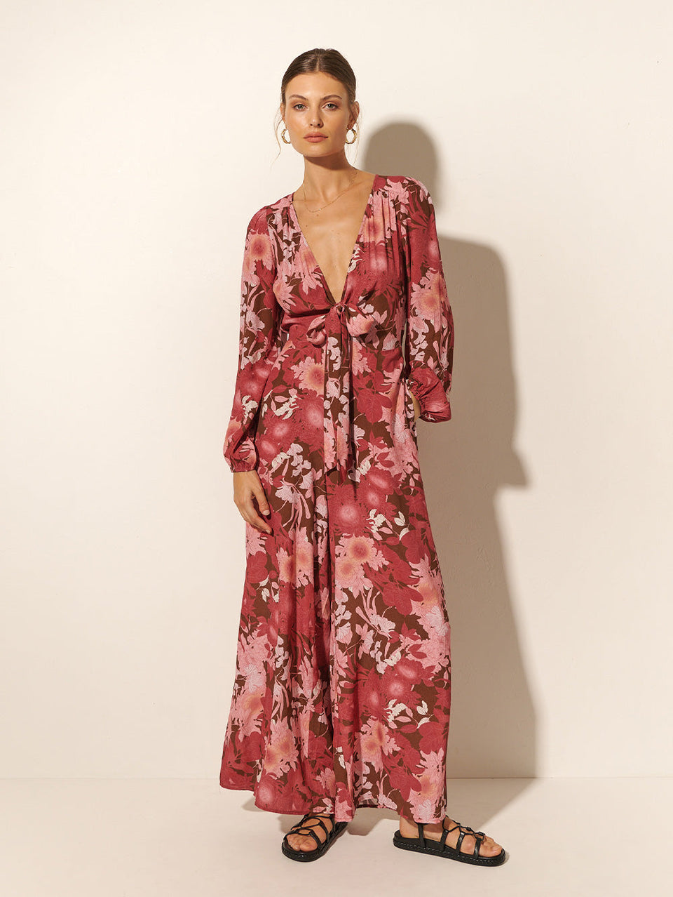 Studio model wears KIVARI Hacienda Jumpsuit: A red, pink and brown floral jumpsuit with ribbon tie front and long sleeves, crafted from sustainable LENZING Viscose Crepe.