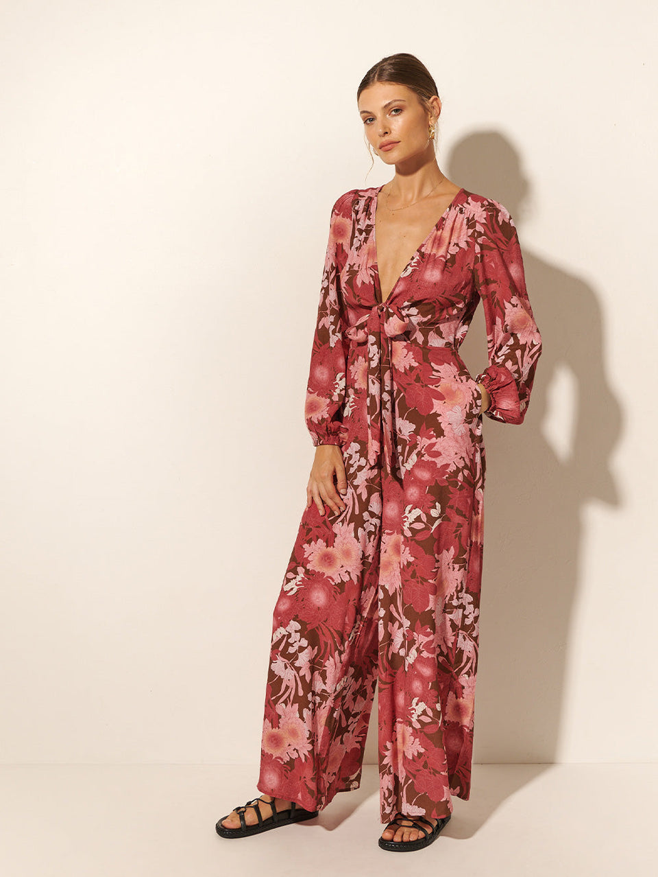 Studio model wears KIVARI Hacienda Jumpsuit: A red, pink and brown floral jumpsuit with ribbon tie front and long sleeves, crafted from sustainable LENZING Viscose Crepe.