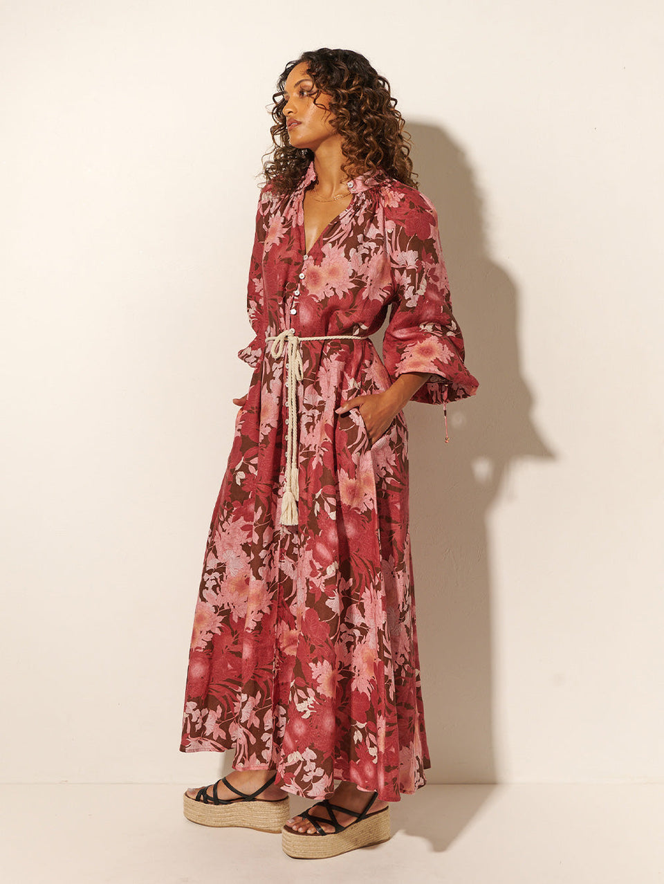 Studio model wears KIVARI Hacienda Maxi Dress: A red, pink and brown floral dress with a shirred collar, button-through front, waist ties and full-length sleeves, crafted from sustainable LENZING Viscose Crepe.