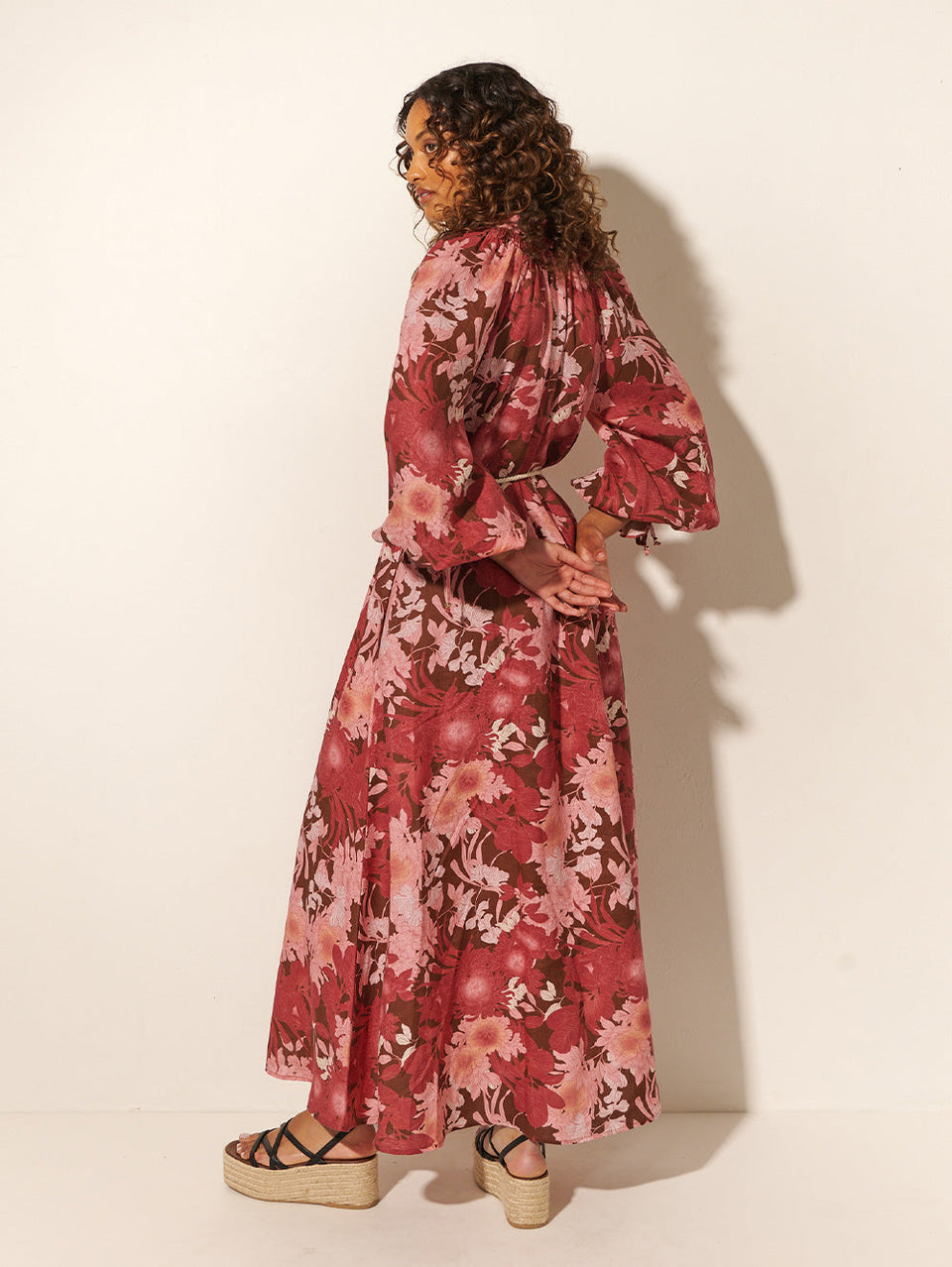 Back shot: Studio model wears KIVARI Hacienda Maxi Dress: A red, pink and brown floral dress with a shirred collar, button-through front, waist ties and full-length sleeves, crafted from sustainable LENZING Viscose Crepe.