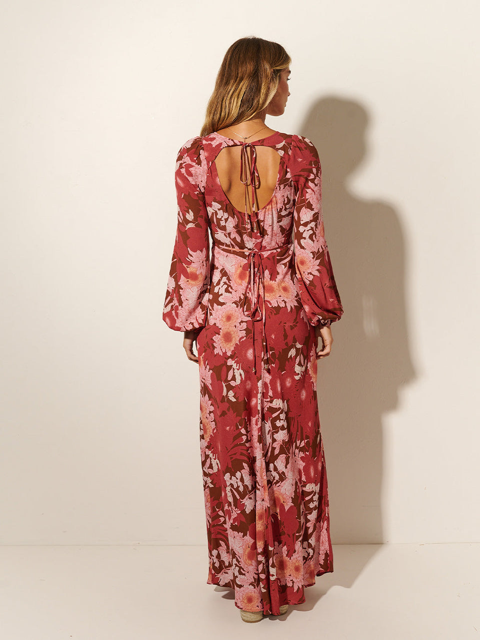 Back shot: Studio model wears KIVARI Hacienda Tie Back Maxi Dress: A red, pink and brown floral dress crafted from sustainable LENZING Viscose Crepe in a bias cut with underbust seams, long sleeves and cut-out back with keyhole ties.