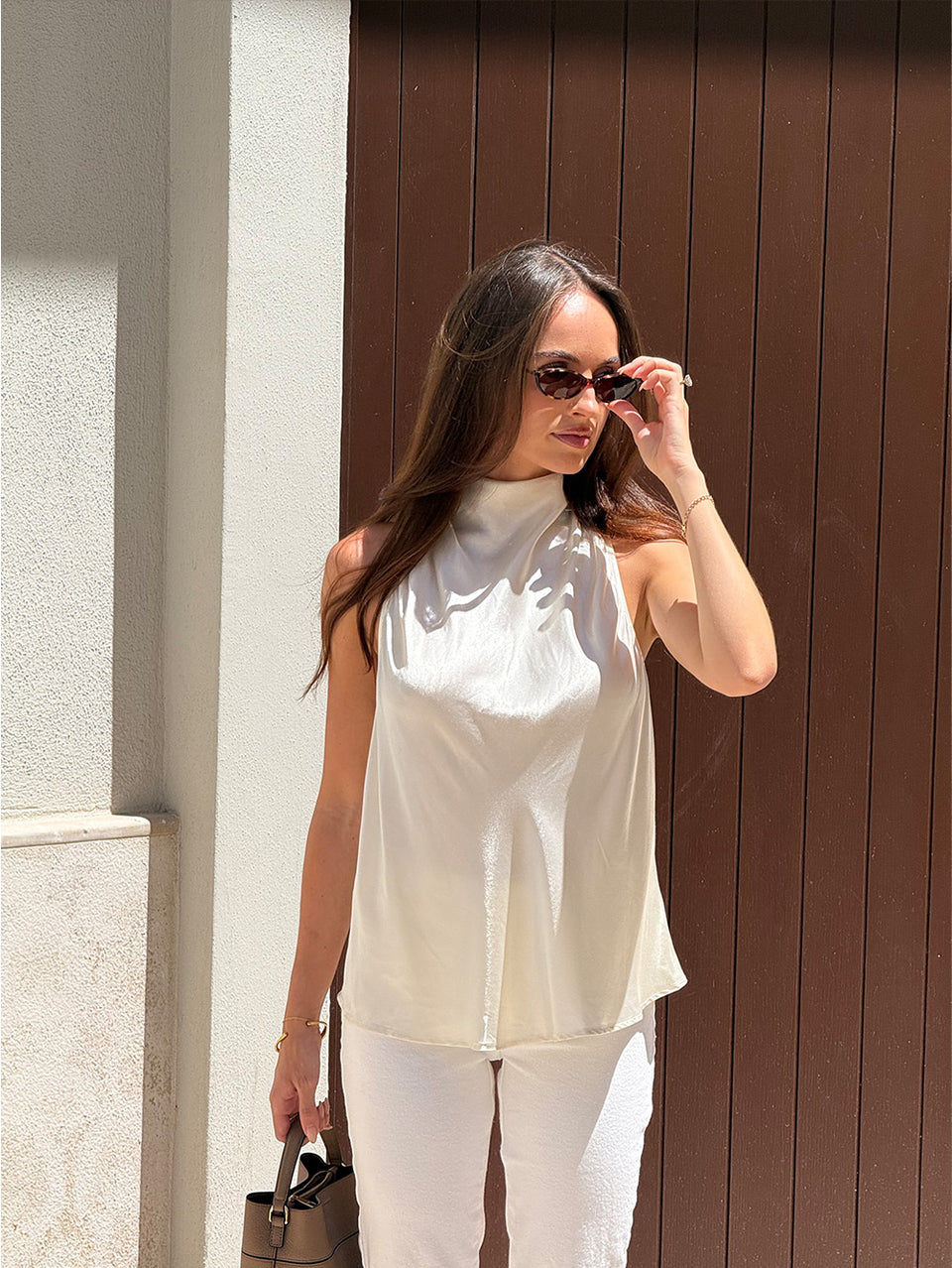 KIVARI Halle Top in White – model wearing women's white sleeveless high-neck draped top with white pants and sunglasses, standing against brown wooden door