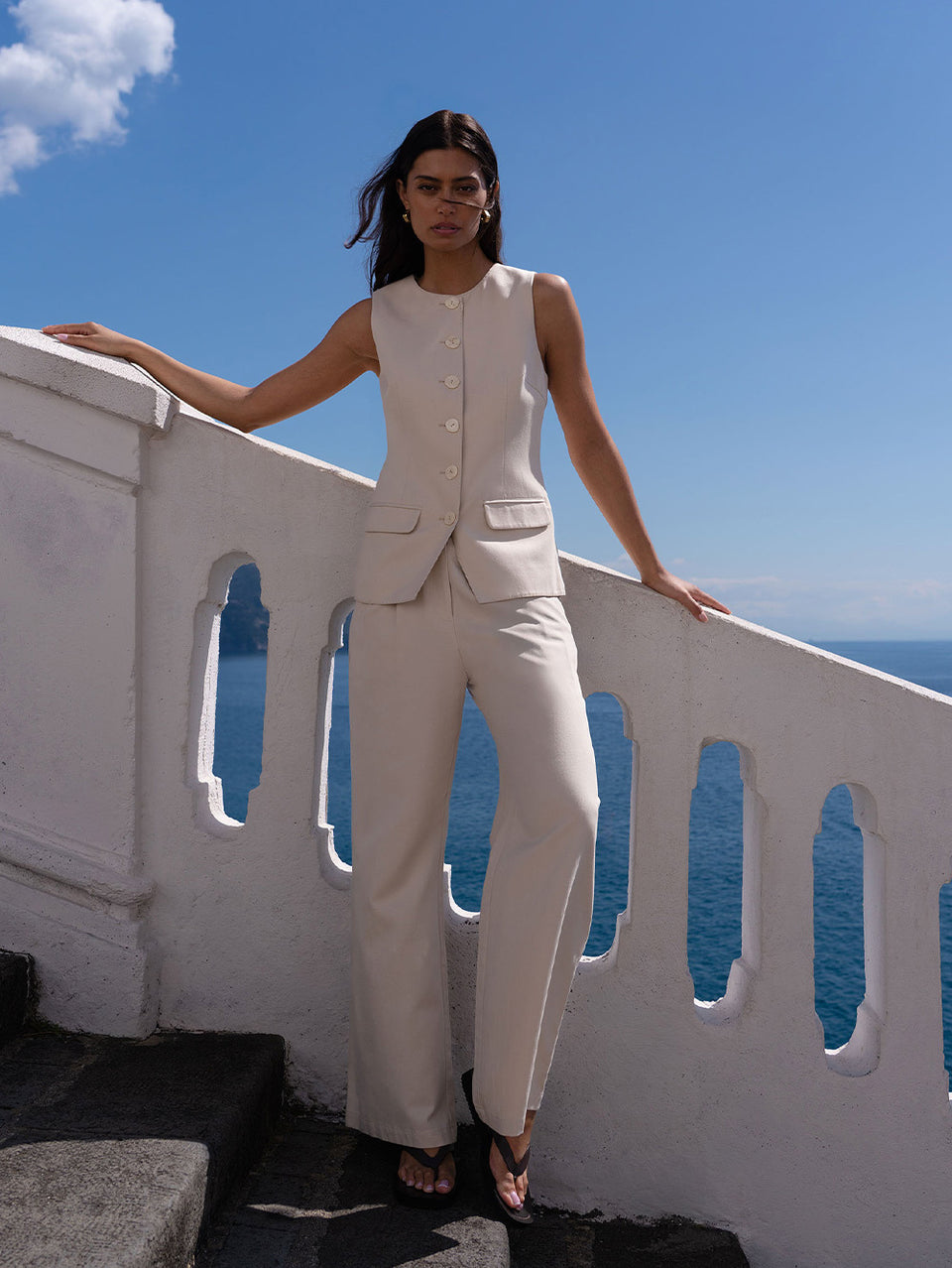 Woman in a beige suit set standing on a balcony with a blue sky and ocean view