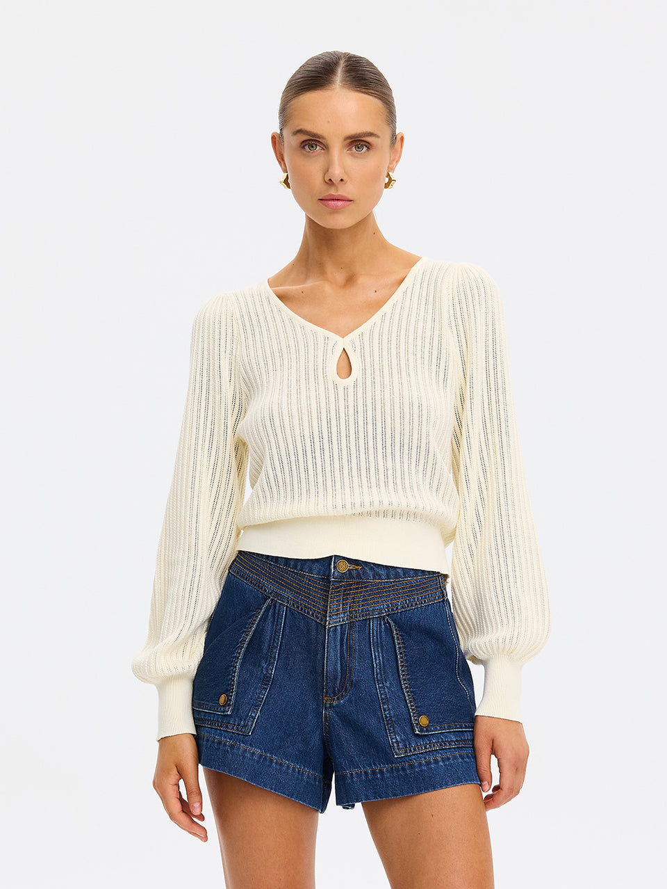 Woman wearing a white ribbed sweater and blue denim shorts on a light gray background