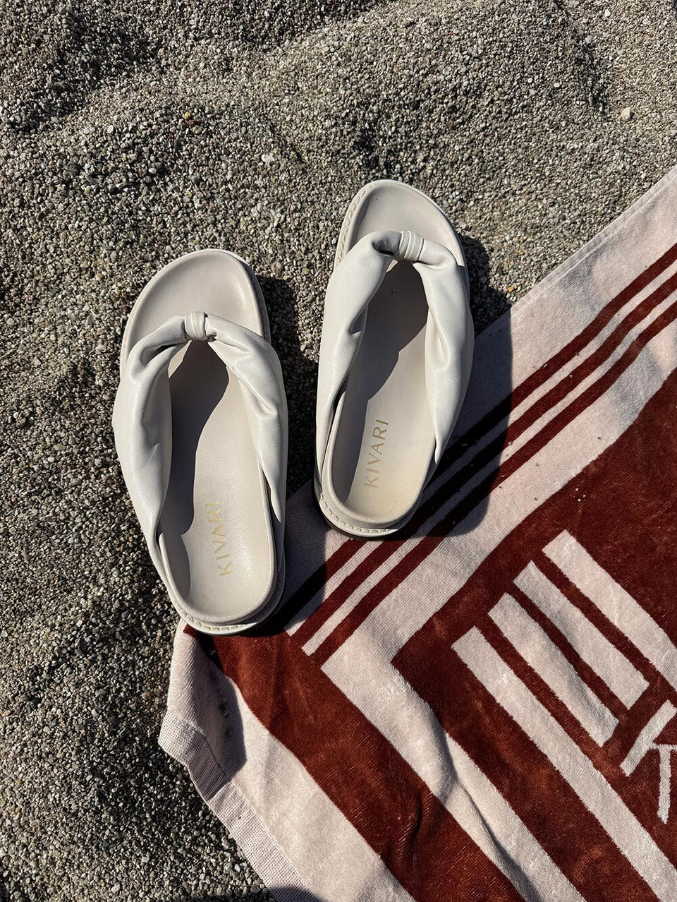 Pair of light creamsandals on a towel with a KIVARI logo on a sandy beach