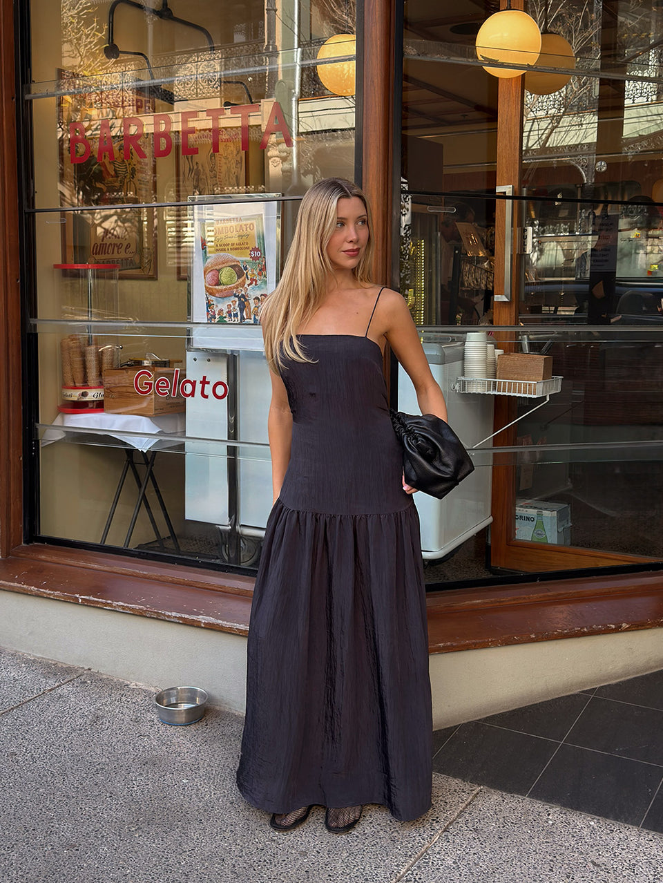 KIVARI Uma Maxi Dress influencer lifestyle shot - model wearing dark navy square-neck spaghetti strap tiered maxi dress holding black bag