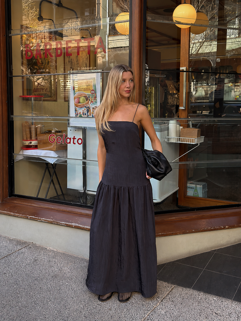 A woman stands on a sidewalk in front of a storefront window wearing a dark charcoal gray maxi dress with thin straps and a gathered skirt. She is holding a black clutch bag.