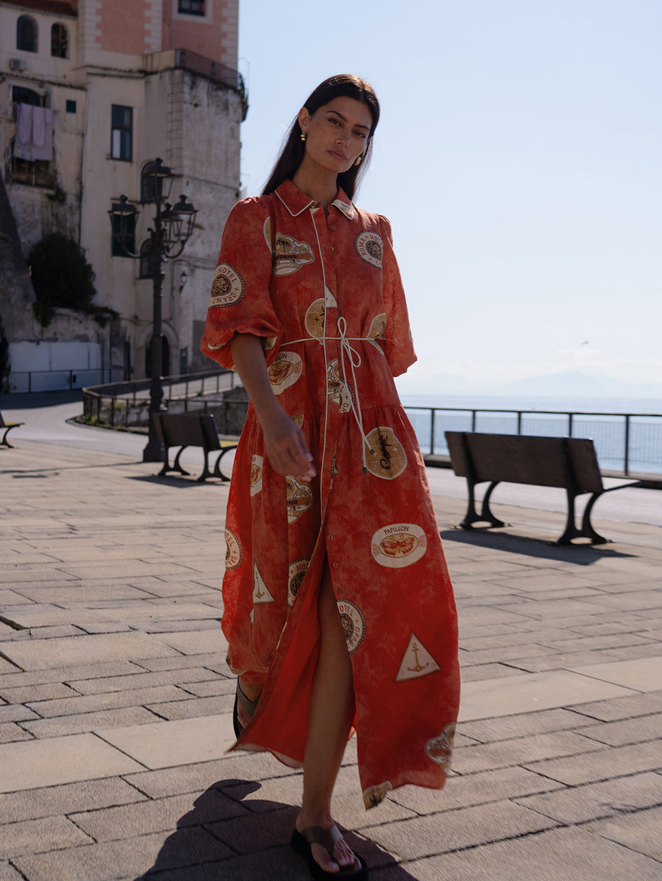 Woman in a red dress with patterns standing on a sunlit street with buildings and benches in the background.