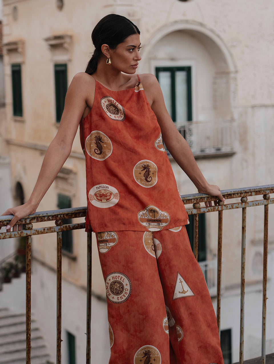Woman wearing an orange patterned outfit standing on a balcony with a building in the background