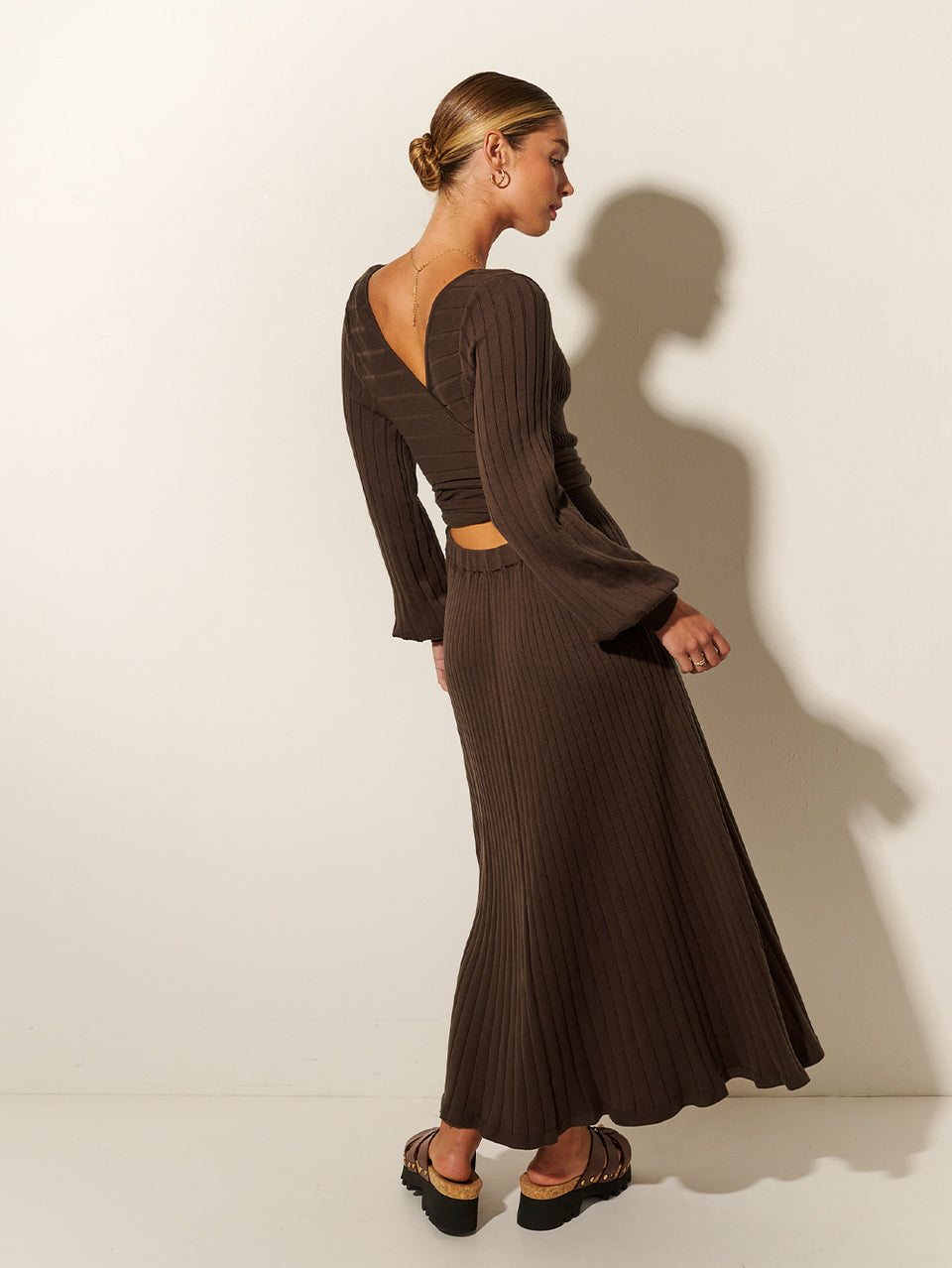 Back shot: Studio model wears the KIVARI Yolanda Long Sleeve Knit Dress - Chocolate: A rich brown reversible dress made from rib knit stitching with soft underbust seams, a tie-back feature with cut-out detail, full-length blouson sleeves and an A-line skirt.